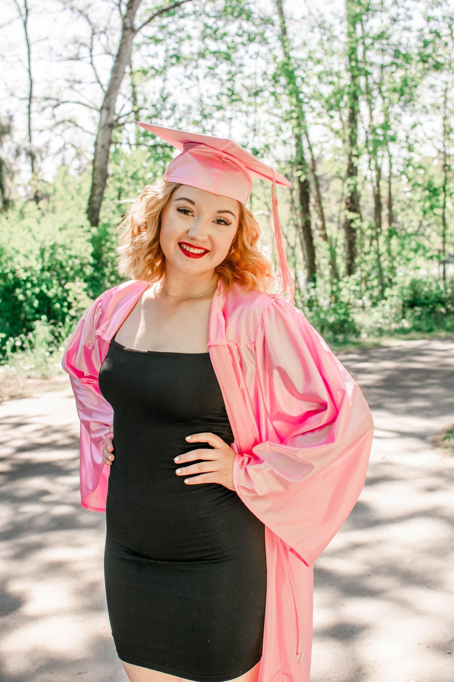 Graduate in black dress and pink graduation gown poses outdoors on wooded path.