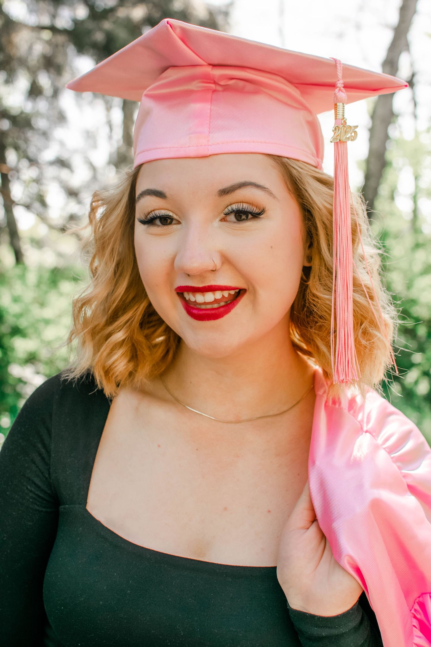 Graduate in pink cap and gown with bright red lipstick smiling outdoors.