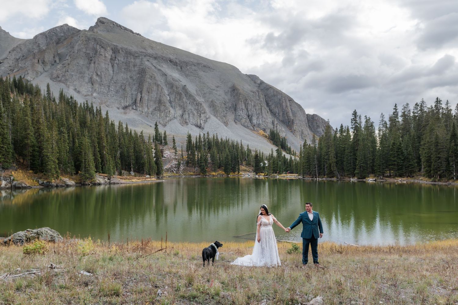 Bride and groom hold hands and walking with their dog by a mountain lake surrounded by evergreen trees and dramatic peaks.