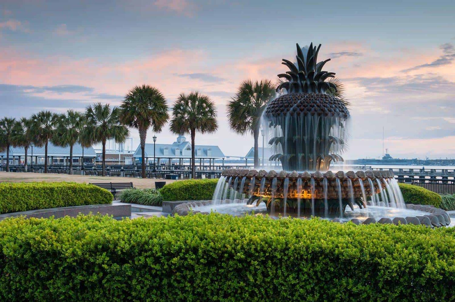 Panoramic sunset view of Charleston Harbor waterfront with palm trees, hedges, and the iconic pineapple fountain.