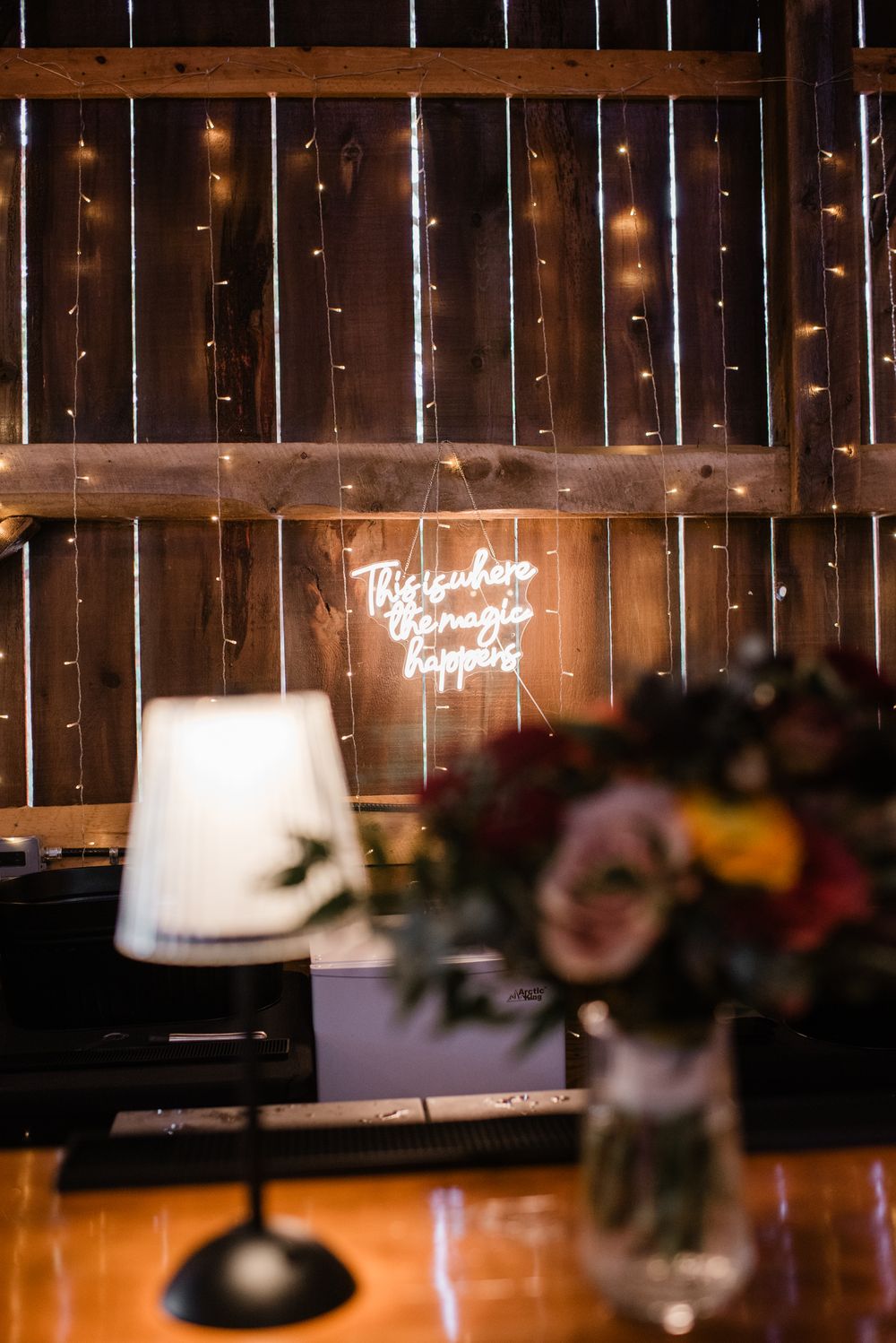 Rustic wooden barn wall decorated with glowing string lights and hanging white lampshades for wedding reception decor.