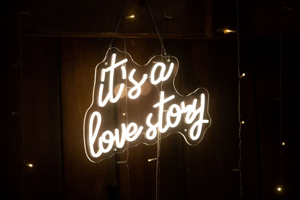 White neon sign reading 'it's a love story' glows against a dark background with small lights scattered around.