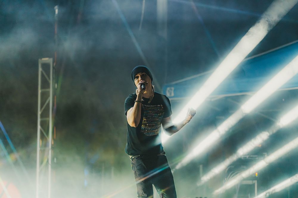 A performer on stage in dark clothing illuminated by bright white spotlights and theatrical fog effects.