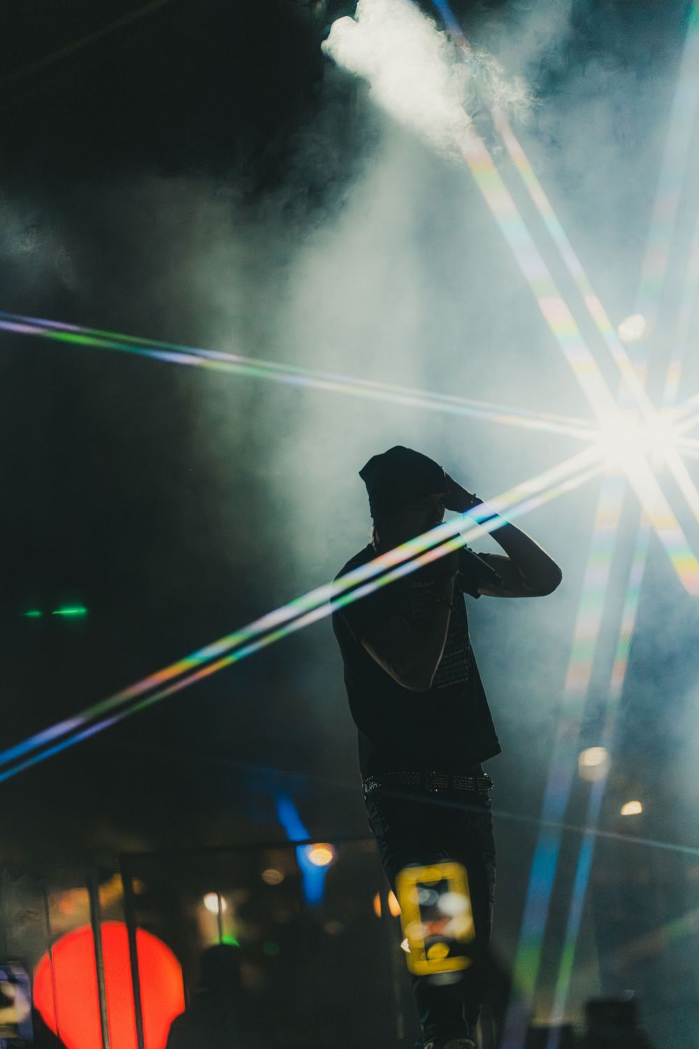 Silhouetted figure stands against dramatic concert lighting with colorful beams and lens flares in dark atmosphere.