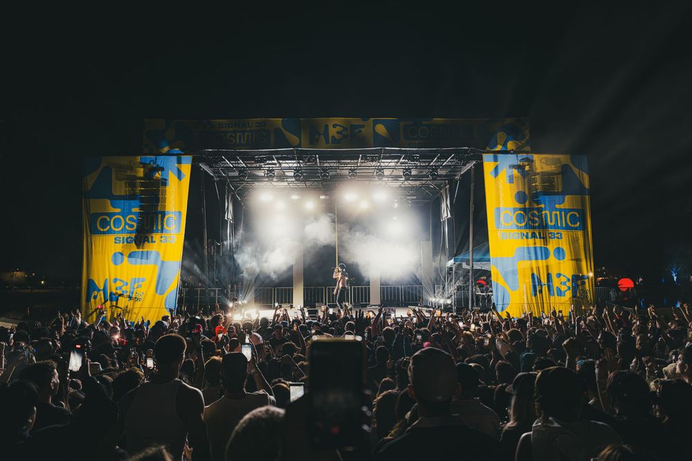 Concert crowd silhouettes in front of illuminated stage with bright yellow banners and atmospheric lighting effects.