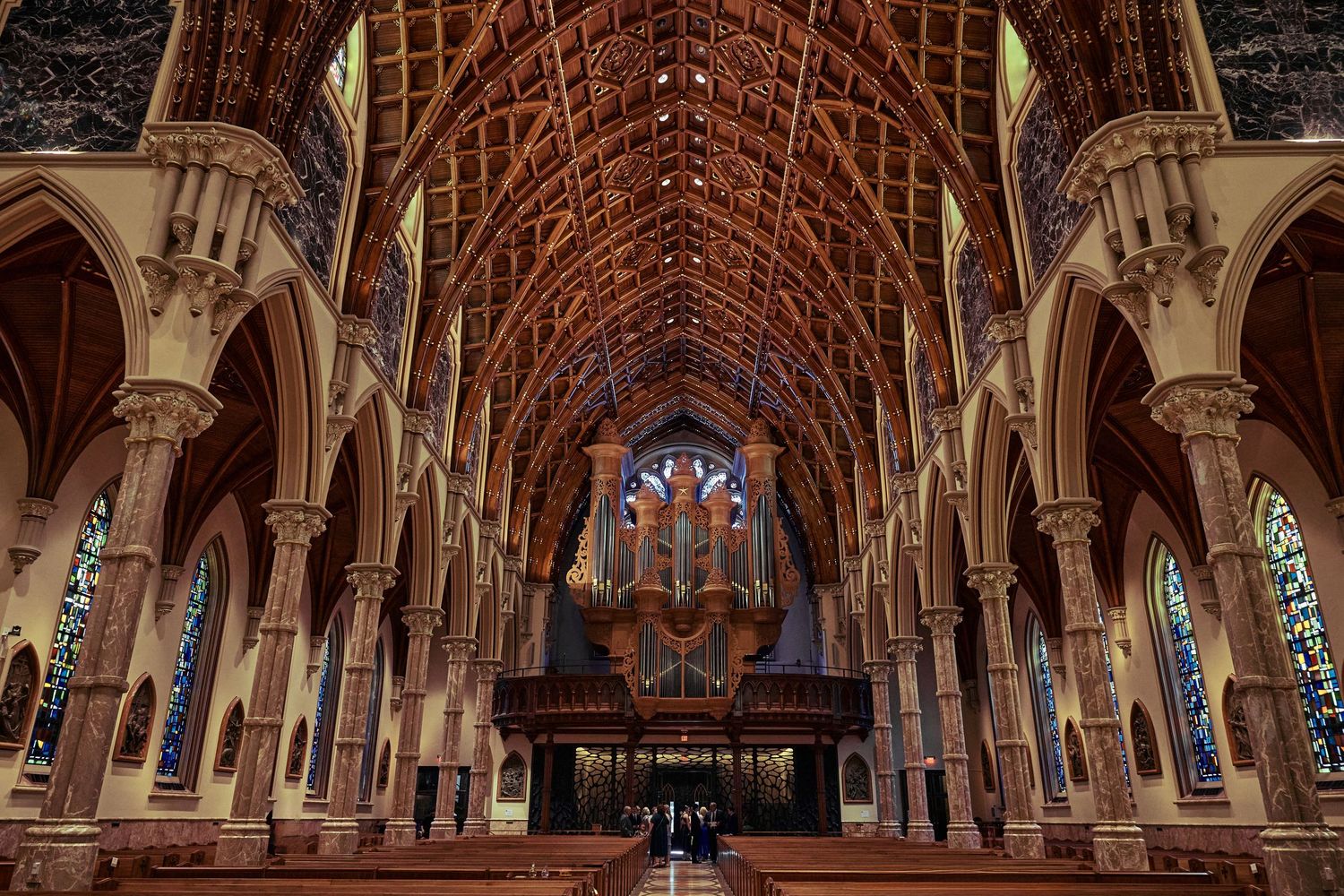 Gothic cathedral interior with ornate wooden ceiling, stone columns, and stained glass windows illuminating the grand space.