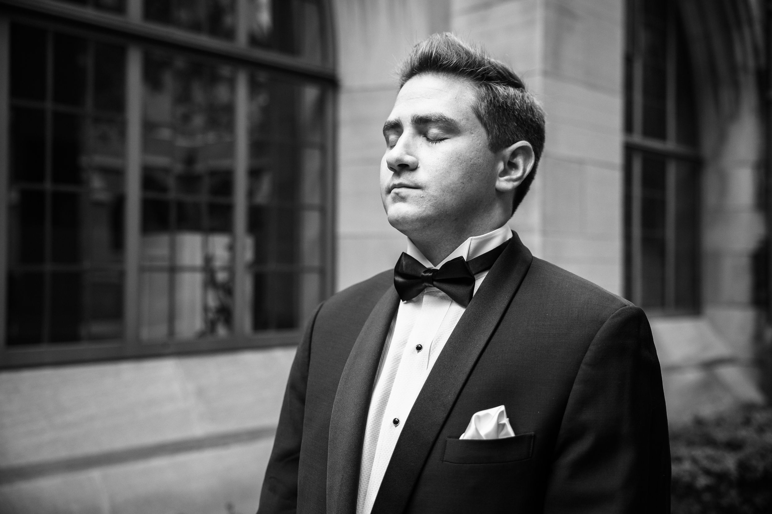 A person in a black tuxedo and bow tie stands in front of a brick wall in a black and white photograph.