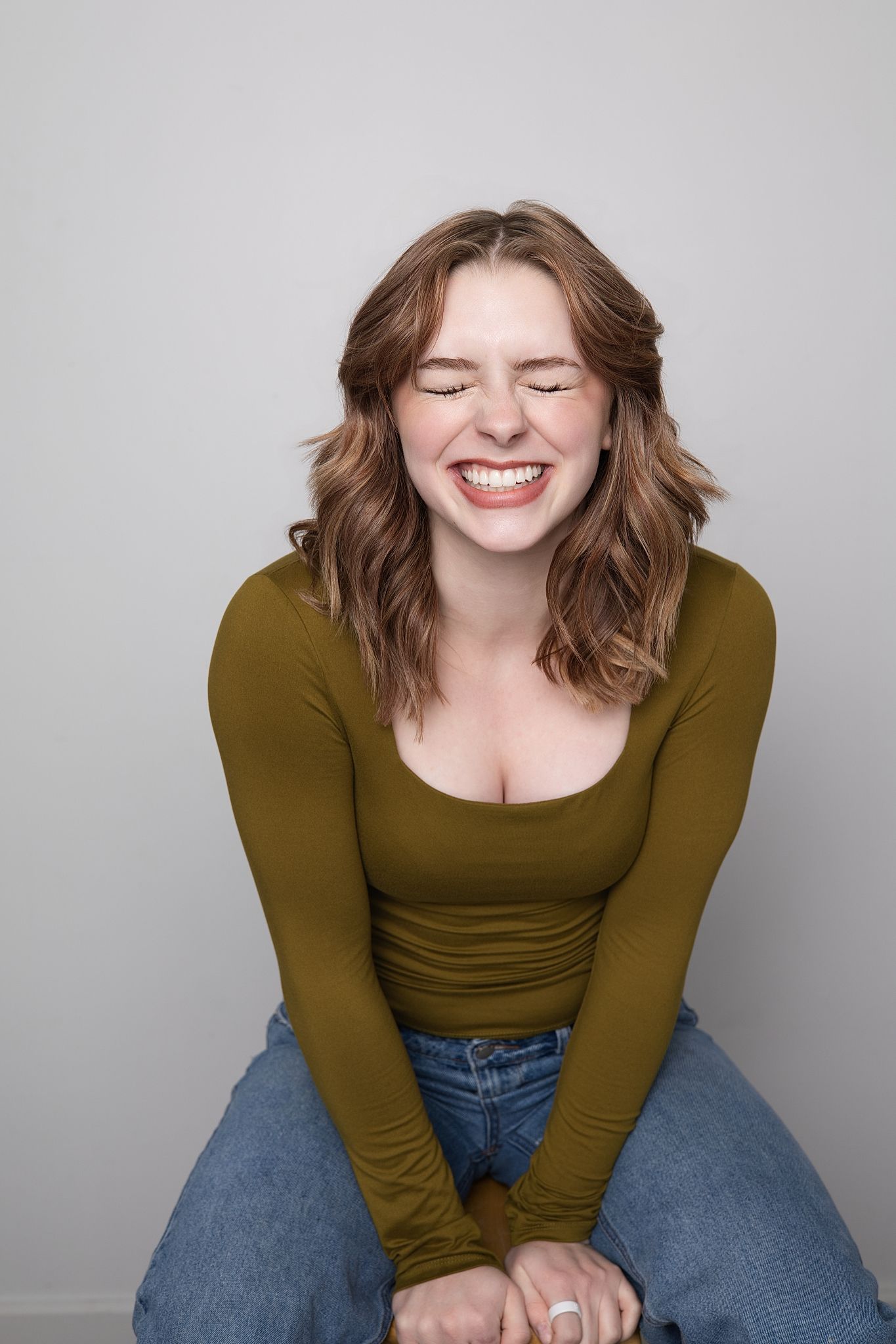 A joyful expression radiates warmth as someone wearing an olive green top and blue jeans sits casually against a gray backdrop.