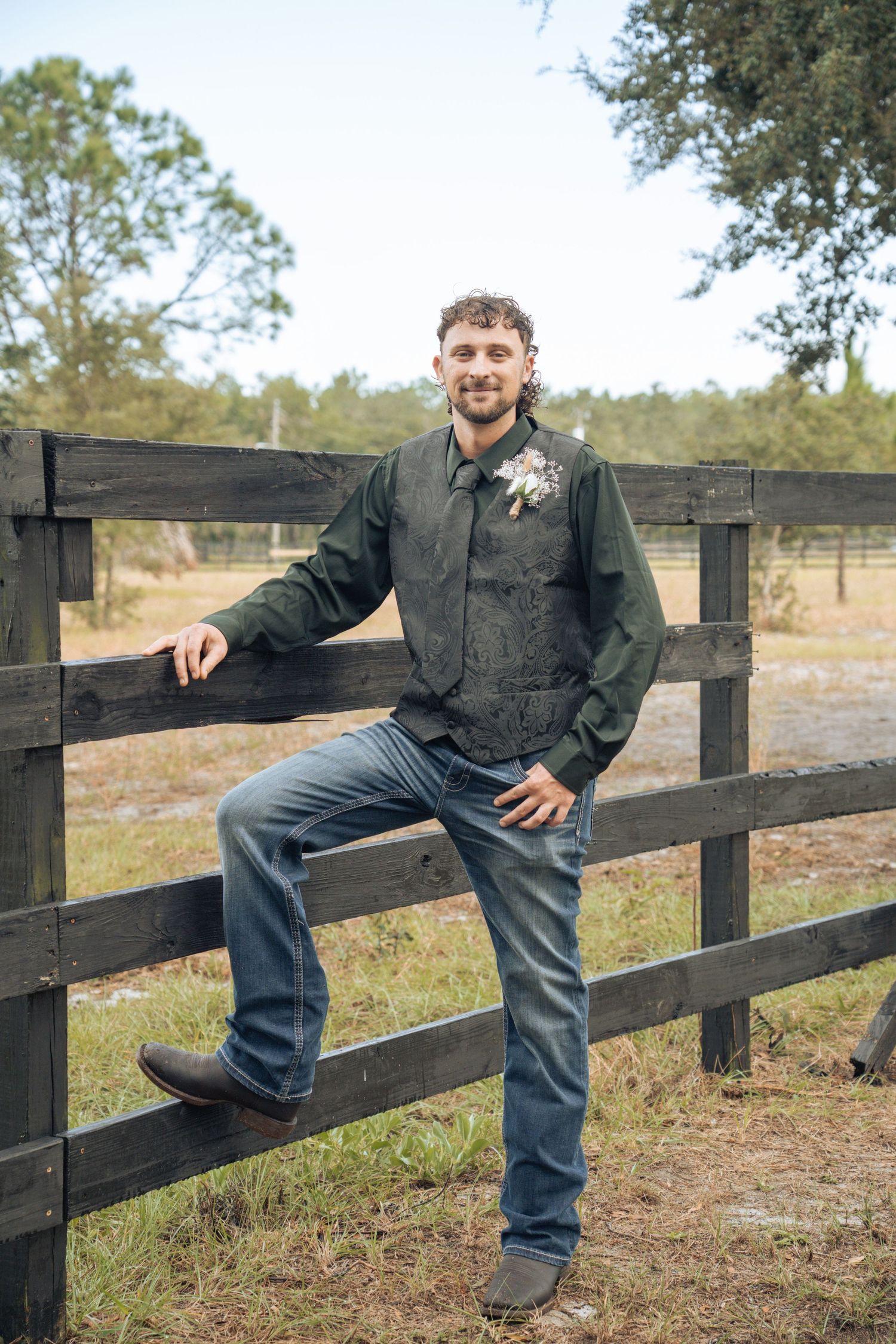 Someone wearing a dark jacket and jeans leans casually against a black wooden fence in a rural outdoor setting.