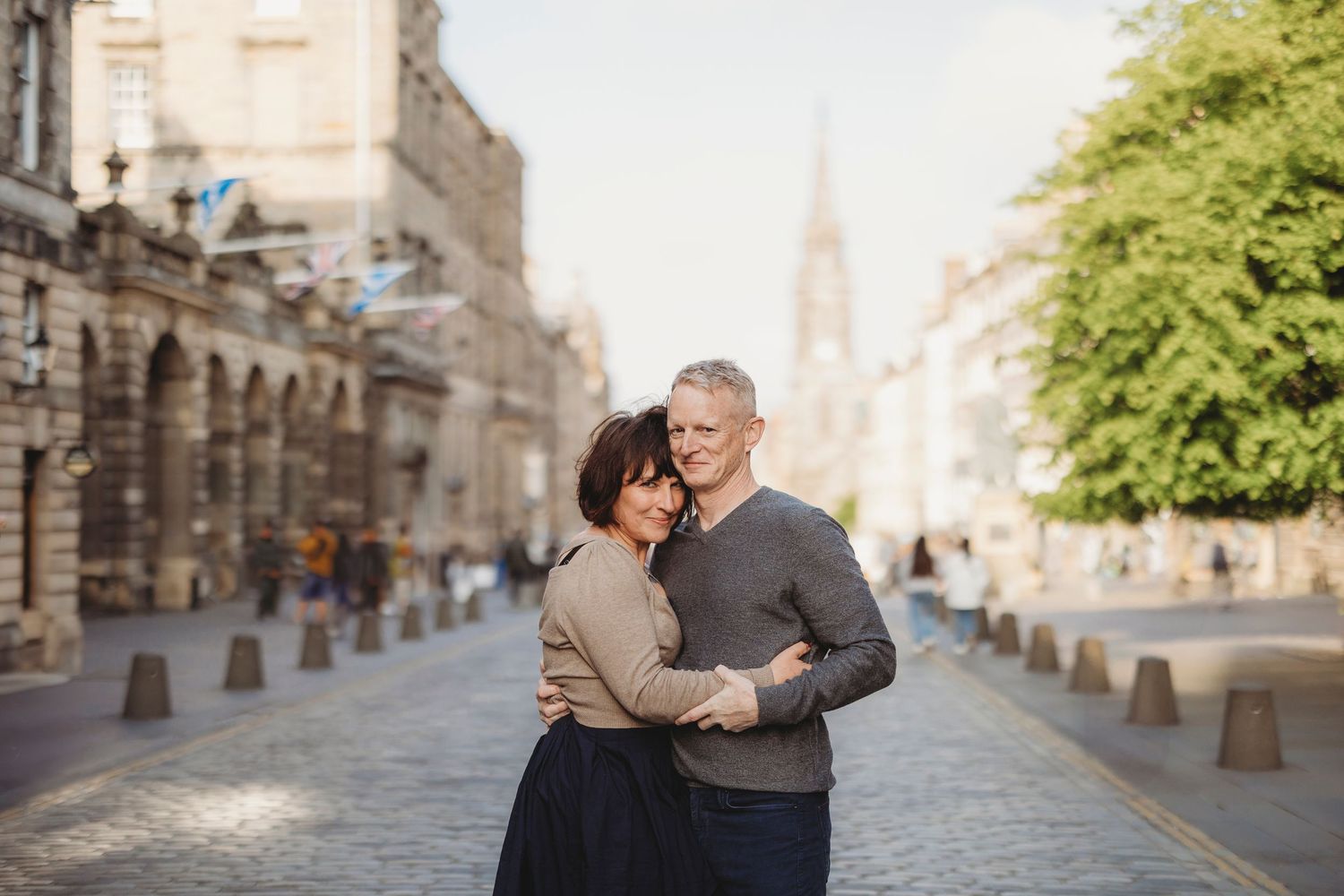 A couple in warm attire shares an intimate moment while dancing on a cobblestone street in a historic European city.
