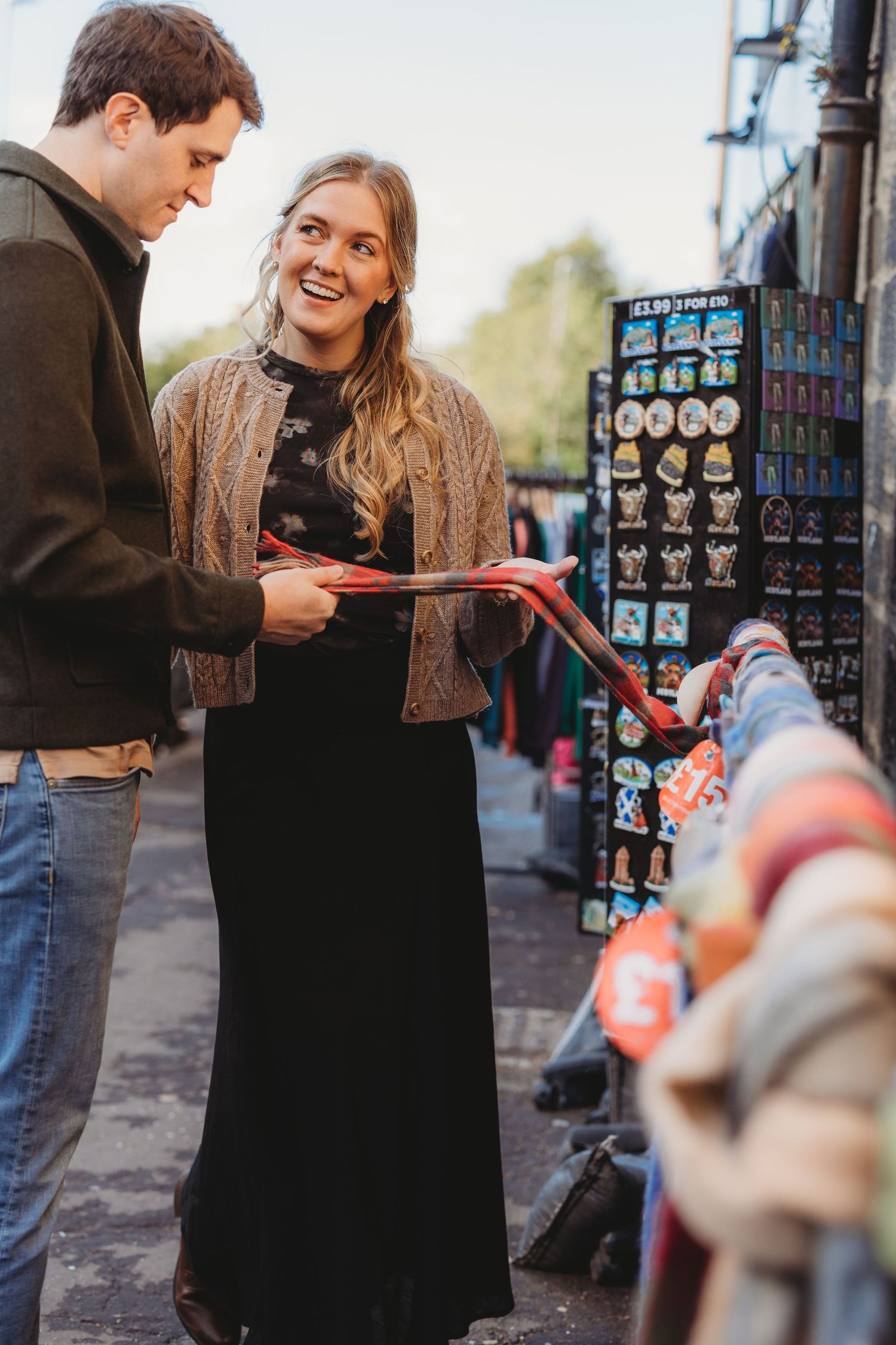 A couple laughs and interacts near a street vendor's display of colorful souvenirs in Edinburgh.