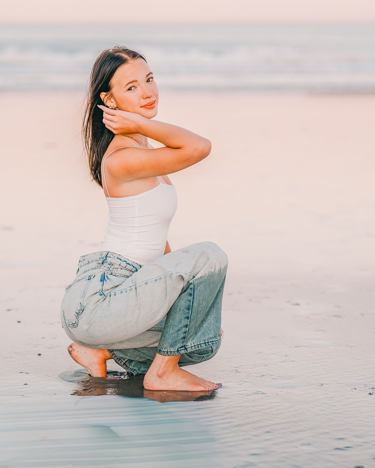 Senior in casual attire crouches on a misty beach at sunset creating a peaceful coastal scene in Hampton Beach, NH