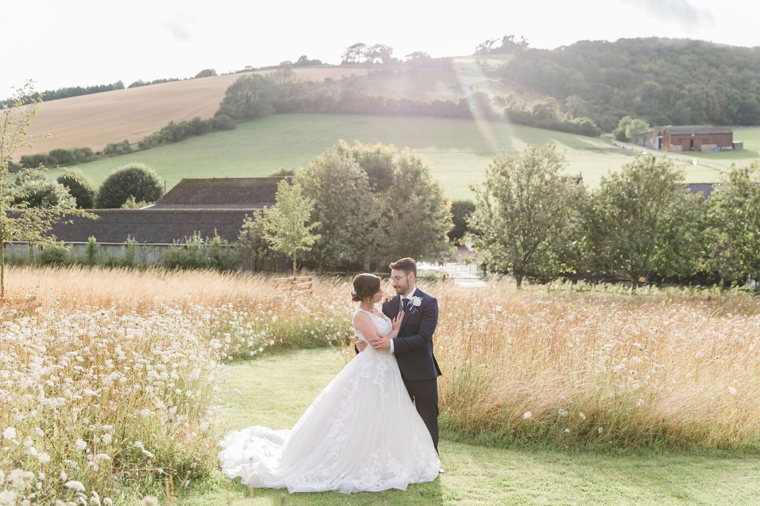 A couple walks through a picturesque meadow with rolling hills and wildflowers on a sunny summer day.