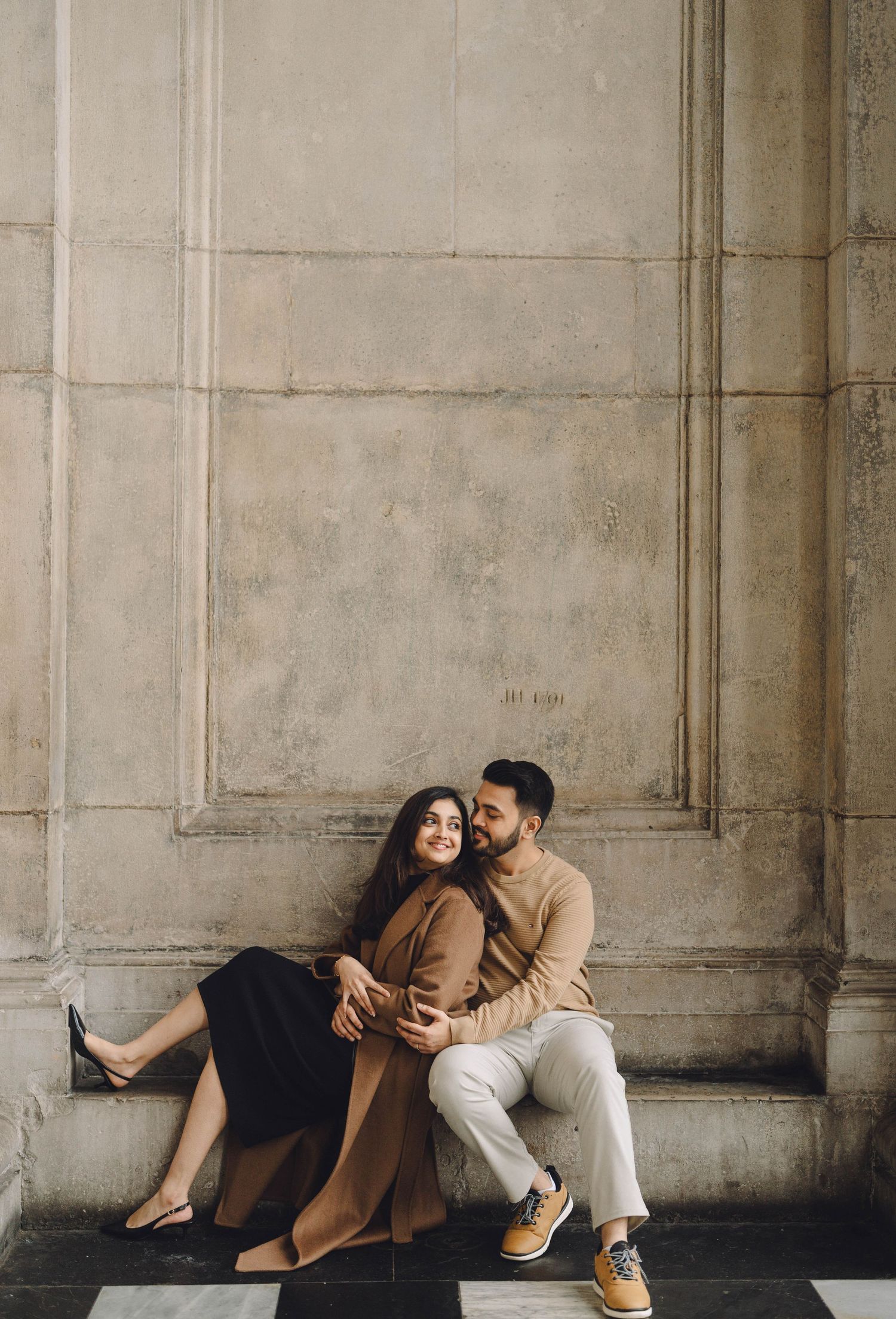 Couple in casual attire sits and leans against stone wall in romantic urban photoshoot.