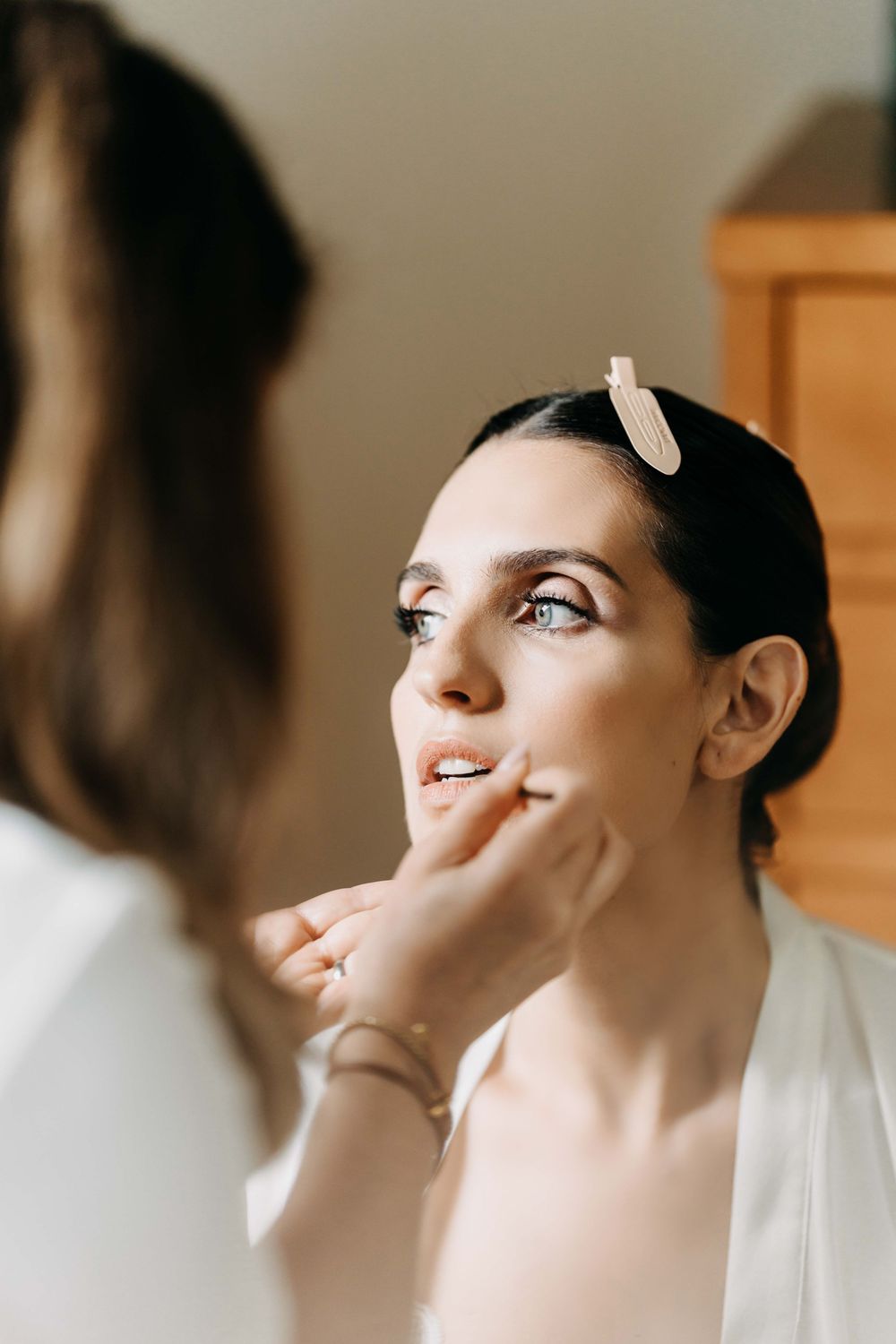 Makeup artist applies lipstick during bridal preparation in soft natural lighting.
