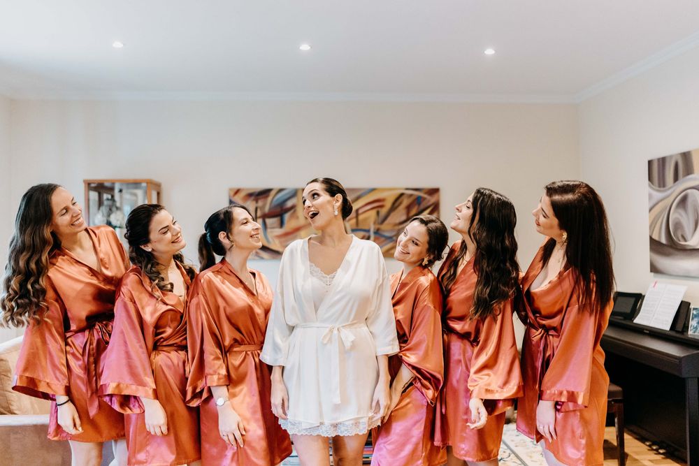 Bridesmaids in coral robes laugh and celebrate together in a bright hotel room before a wedding ceremony.