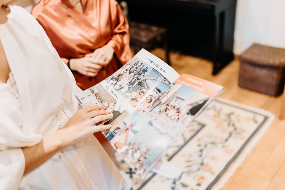A person reads a picture book while sitting at a wooden table with several other books spread out nearby.