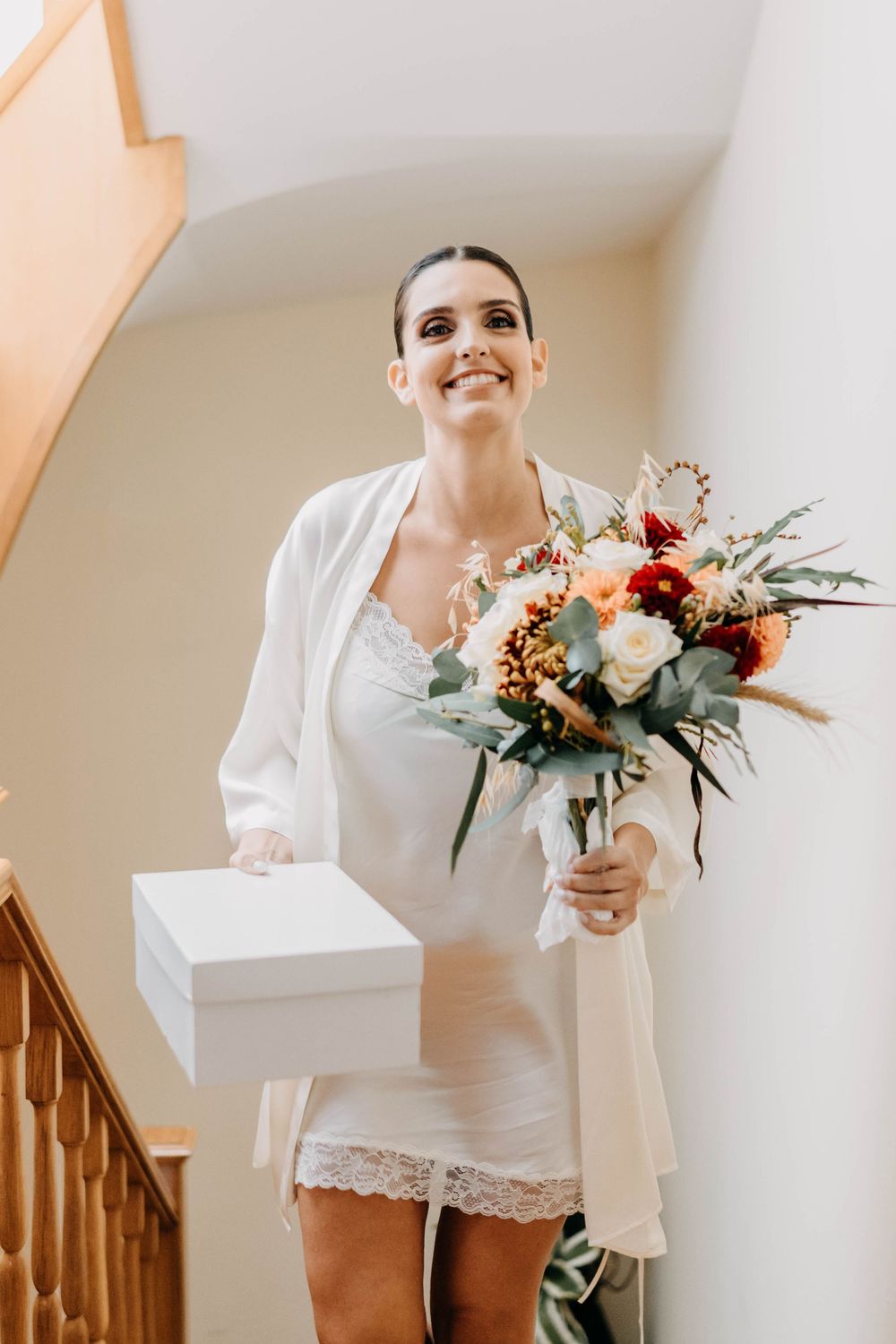 A bride in a short white dress holds a colorful bouquet while standing next to a wooden staircase.