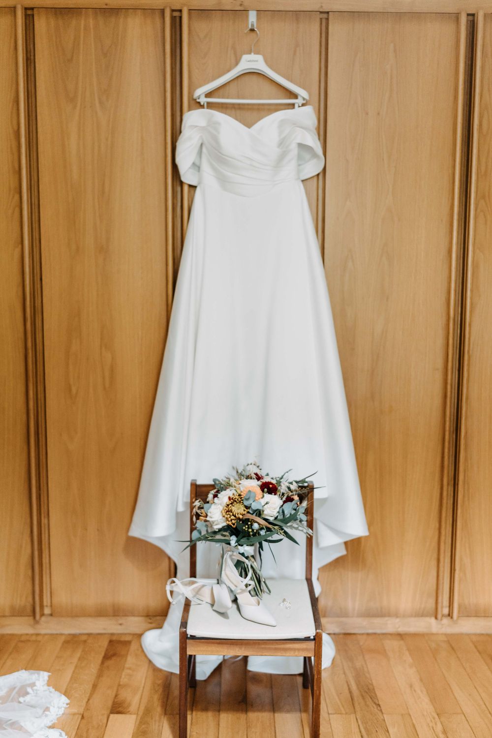 Three elegant white wedding dresses hang on wooden hangers against a light wood paneled wall with floral accents.