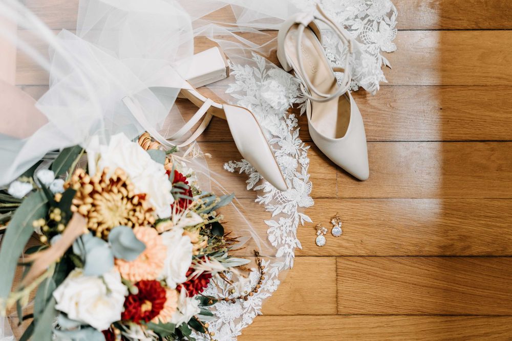 Wedding details with elegant white heels, delicate lace veil, and a romantic bouquet with roses on wooden flooring.