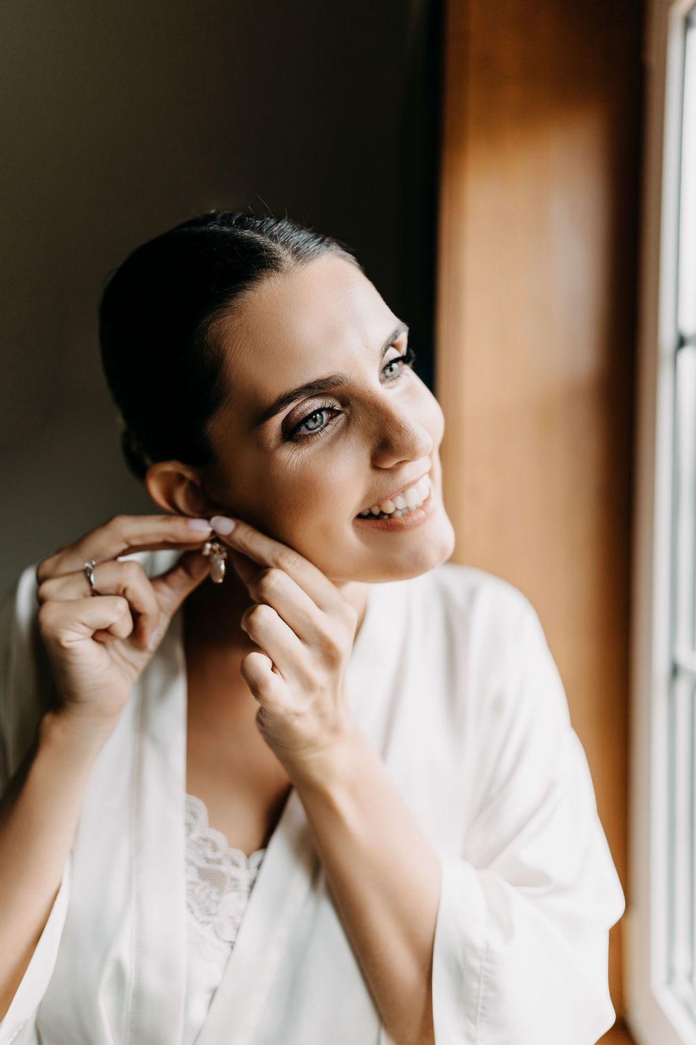 A person in a white robe smiles while putting on earrings near a window with natural lighting.