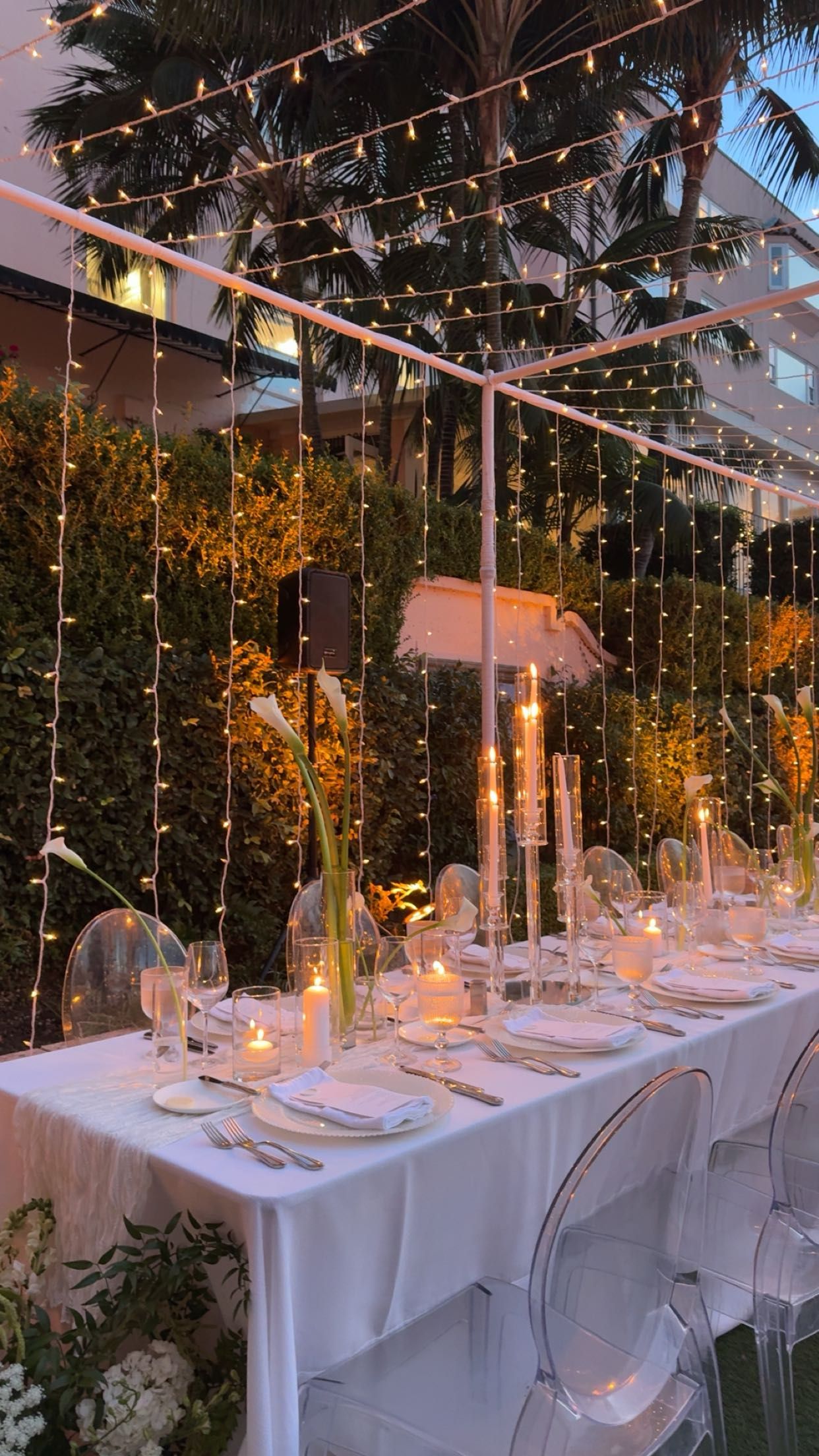 Elegant wedding reception tables set with white linens and clear chairs under cascading string lights at sunset.