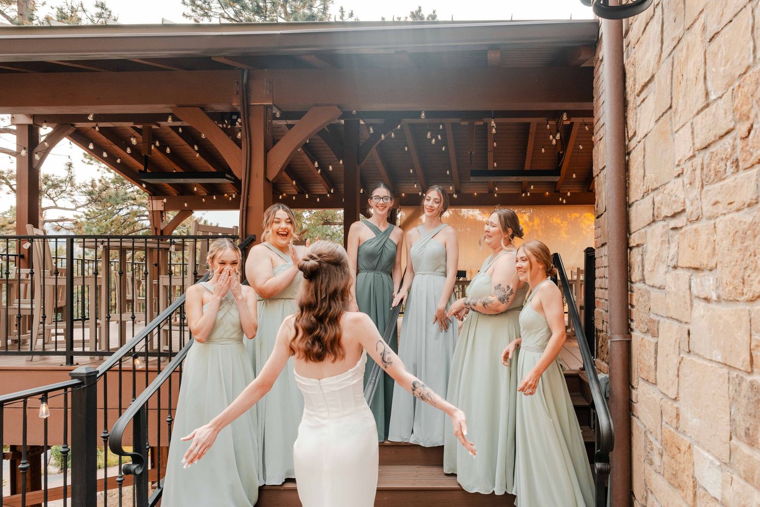 Bride revealing wedding dress to excited bridesmaids in sage green gowns at outdoor venue.