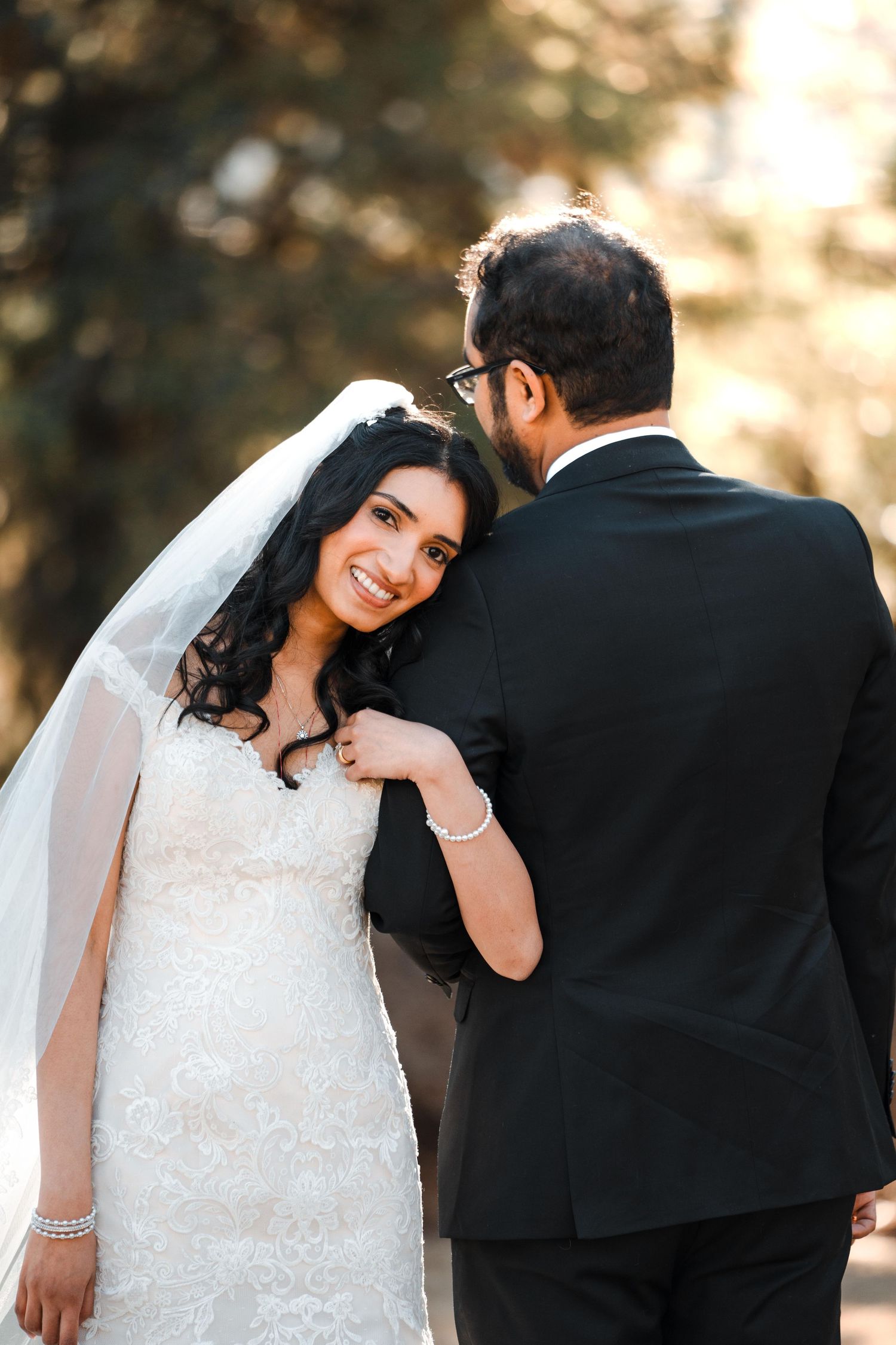 Smiling bride in lace gown leans on groom in black suit during outdoor wedding portrait.