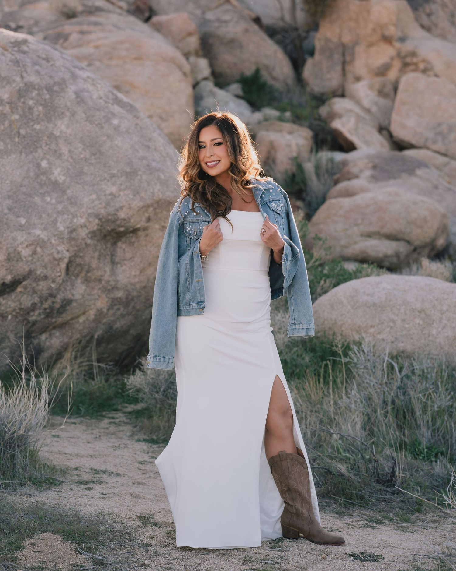 Woman in white maxi dress, denim jacket, and brown cowboy boots posing among large rocks in a desert landscape.