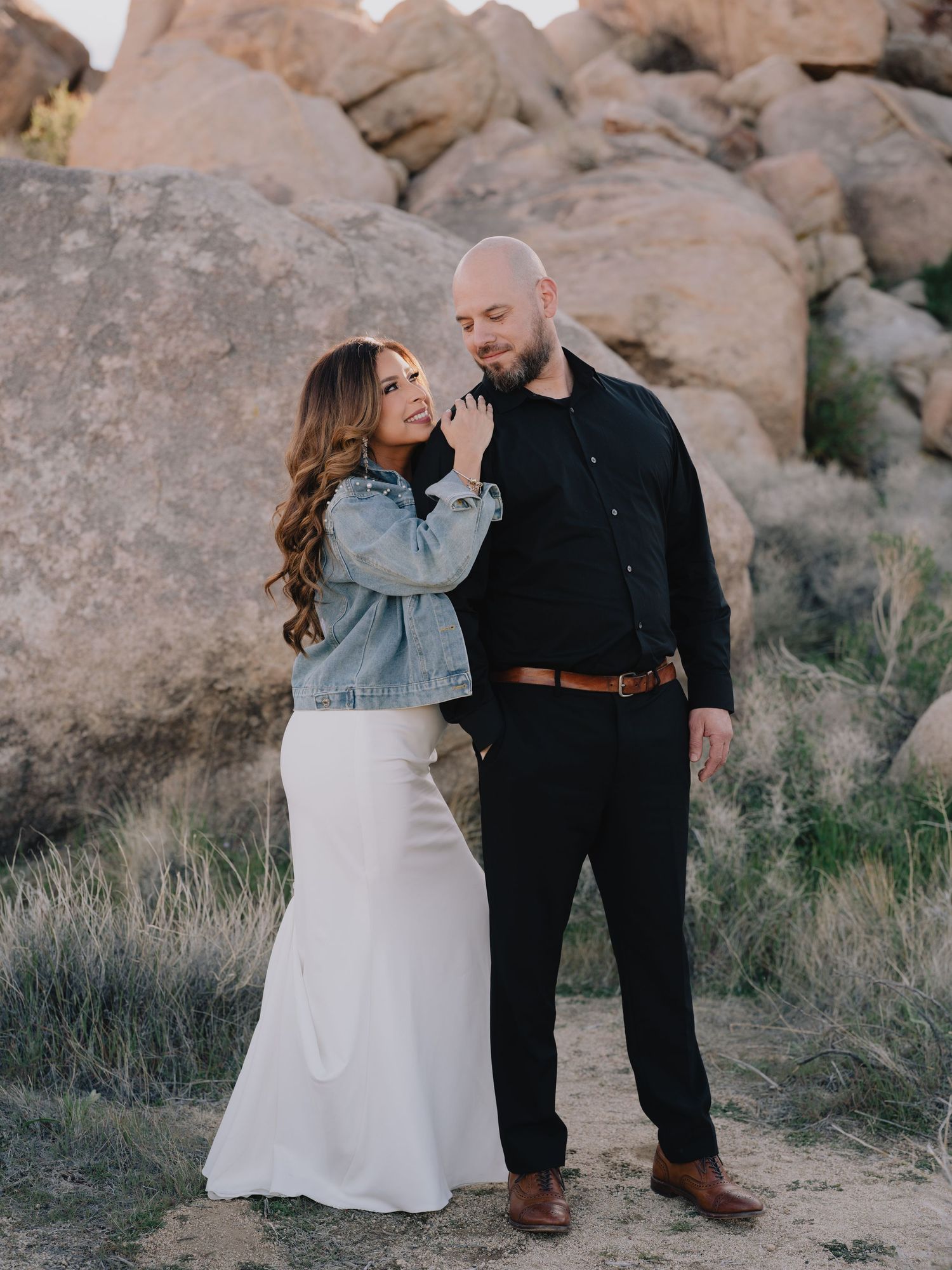Couple posing romantically among desert boulders, woman in white skirt and denim jacket, man in black outfit.