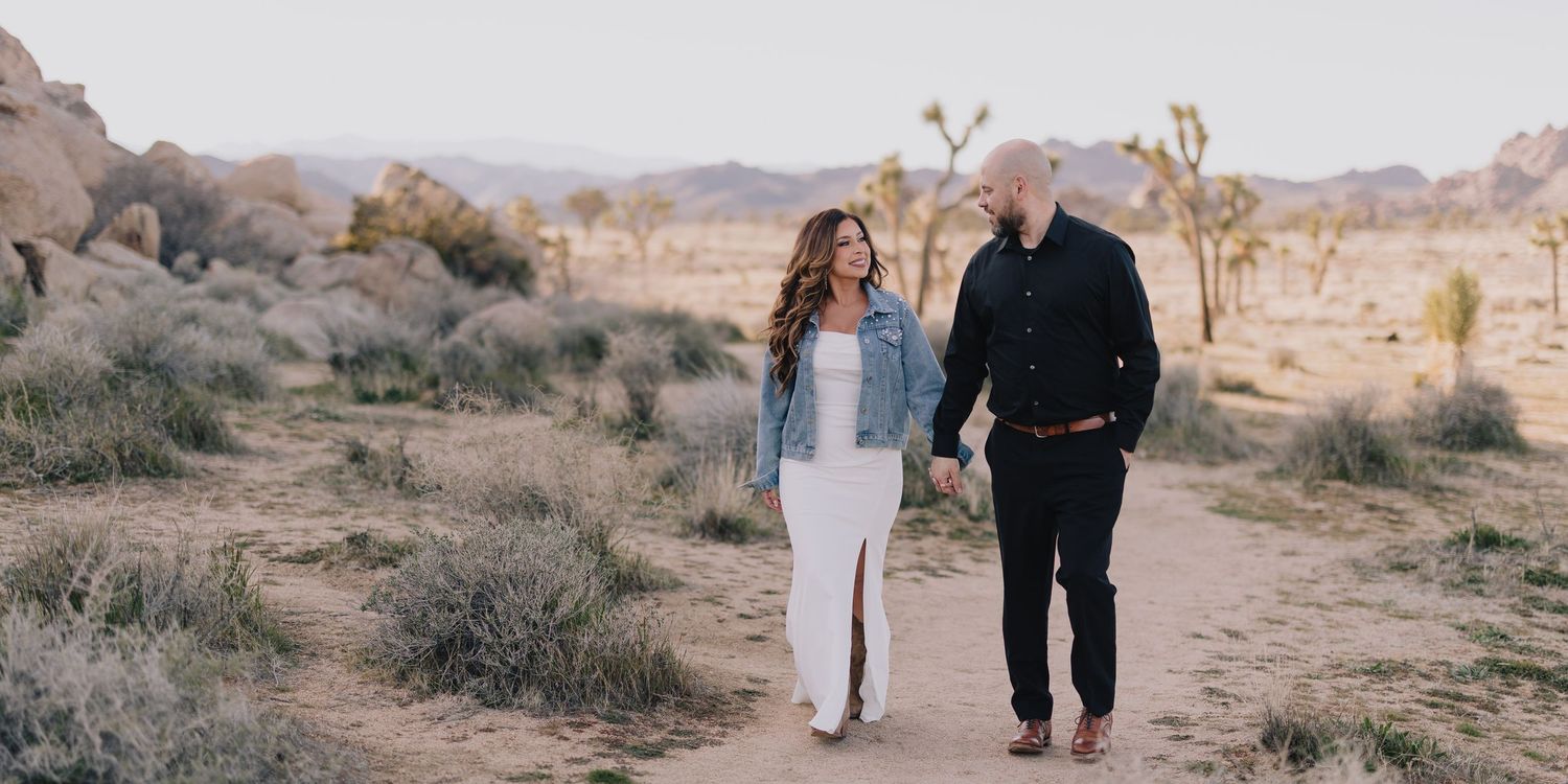 Couple holding hands walking on a desert path in Joshua Tree with rocky landscape and cacti in background.