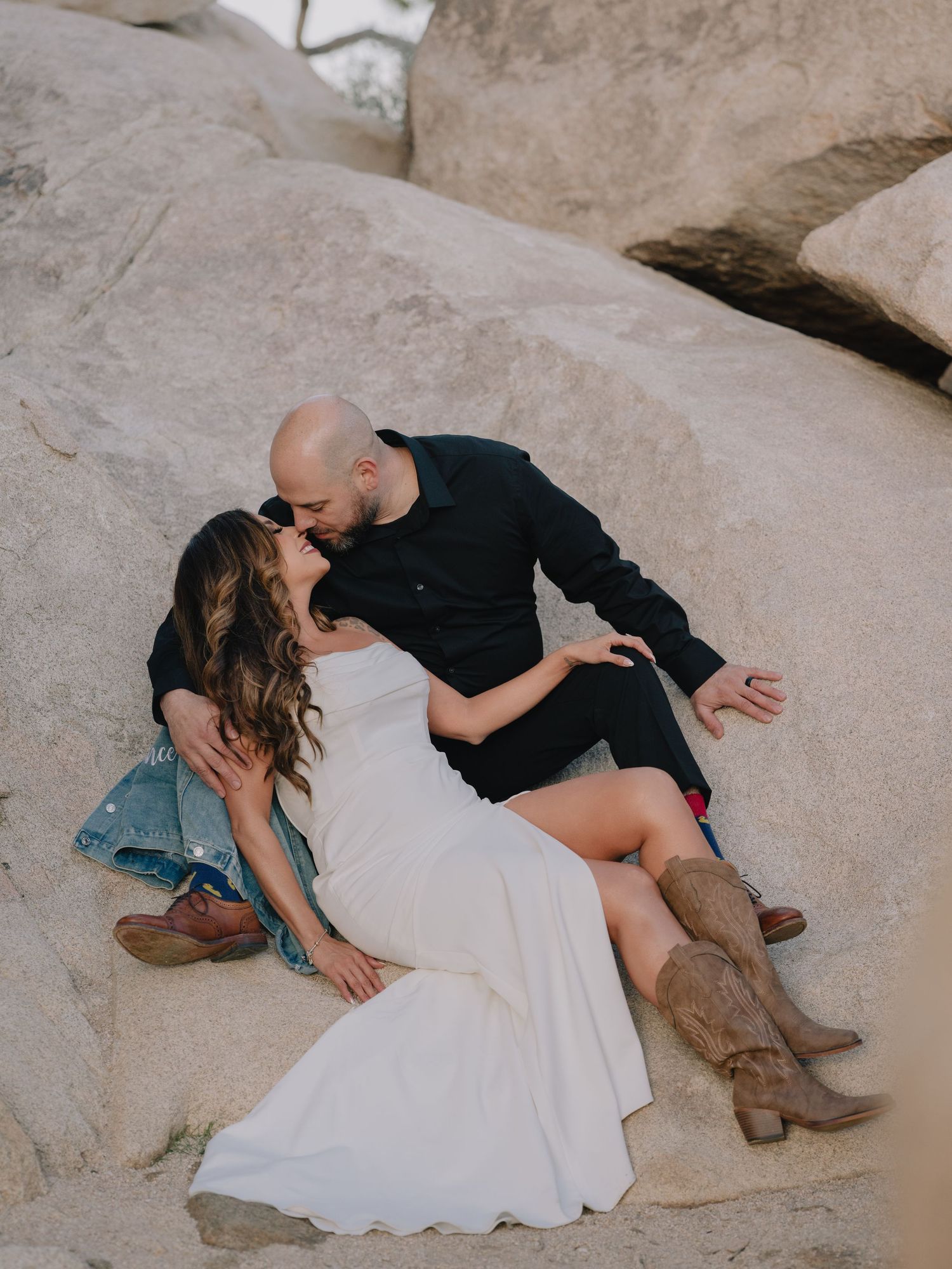 Couple sharing a romantic kiss while sitting on large boulders, bride in white dress and cowboy boots, groom in black outfit.