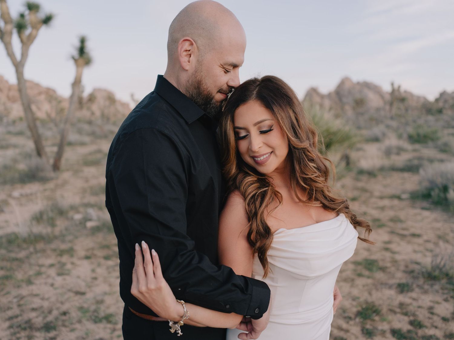 Couple embracing in Joshua Tree desert, man in black shirt holding woman in white off-shoulder dress at dusk.
