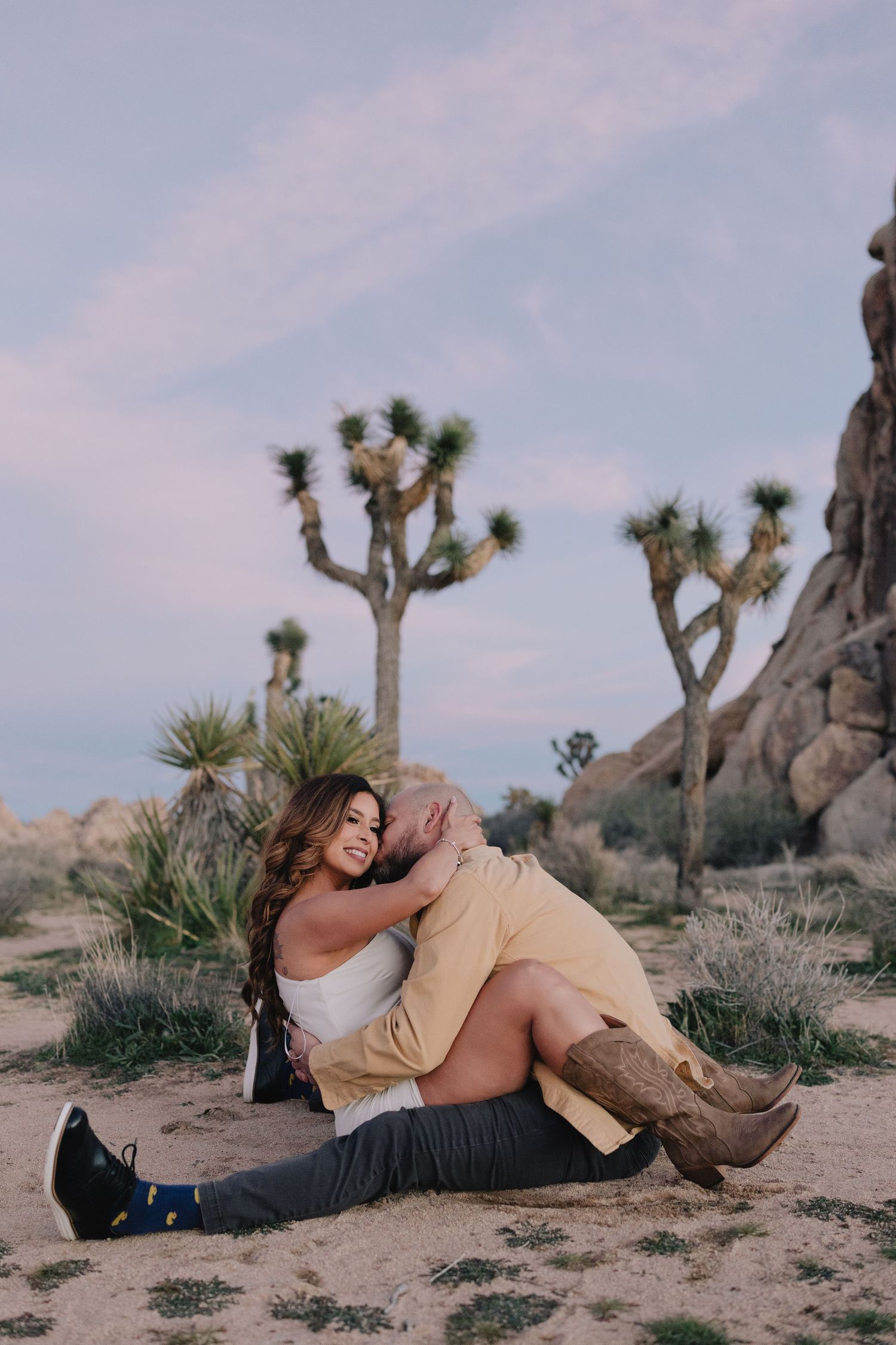 Couple embracing and sitting on desert sand with Joshua trees and rocky formations in the background.