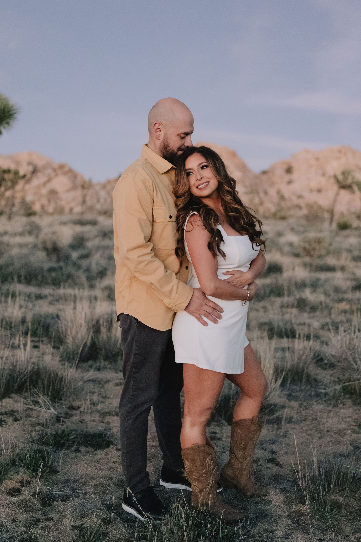 Couple embracing in desert landscape, woman in white dress and cowboy boots, man in yellow shirt, rocky hills background.