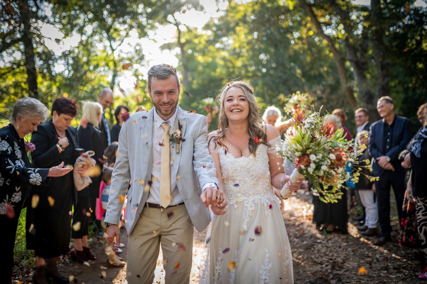 Bride and groom walk hand-in-hand through confetti shower outdoors, surrounded by smiling guests in a sunlit woodland setting.