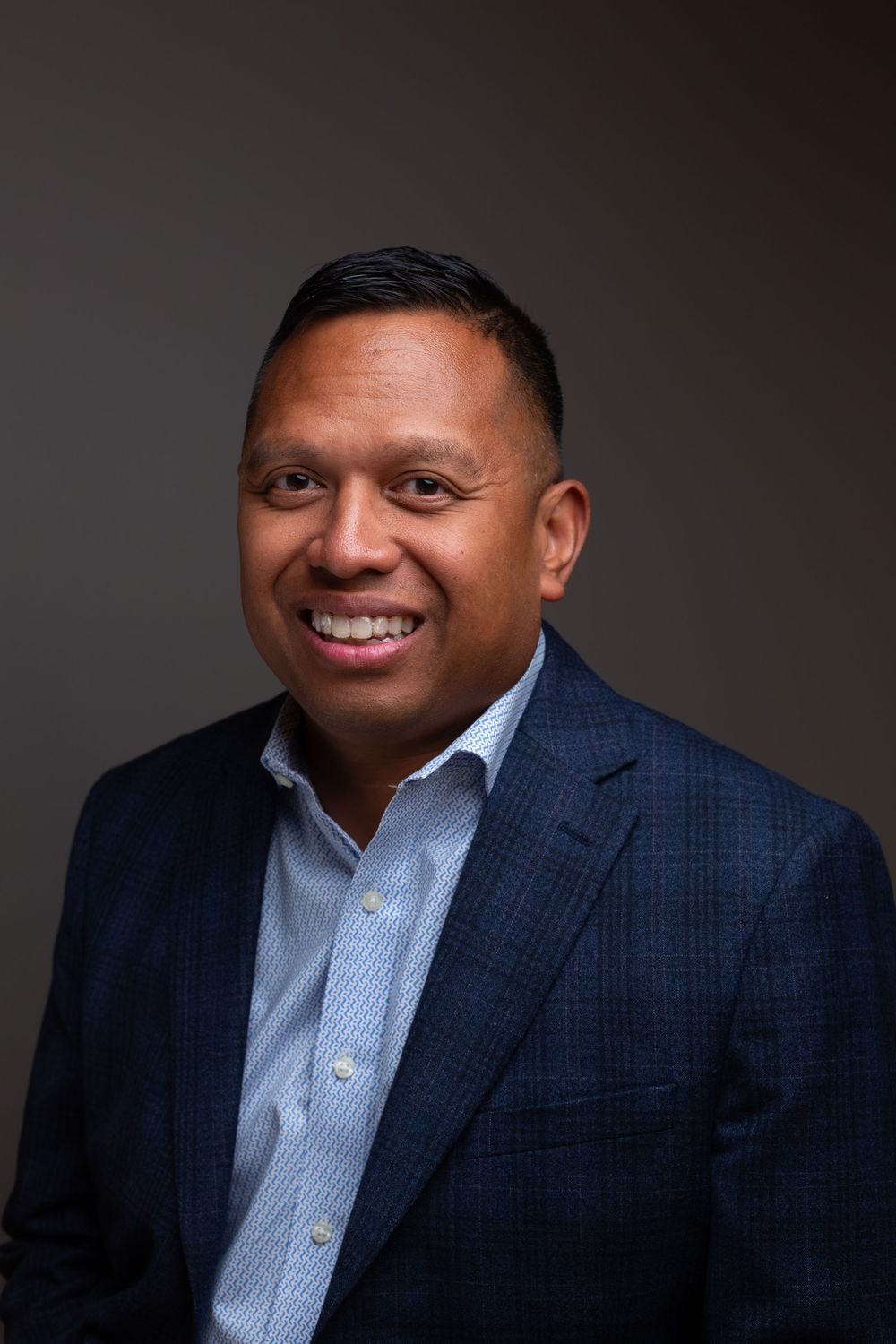 Professional headshot of a smiling man in a navy plaid blazer and light blue shirt against a gray background.
