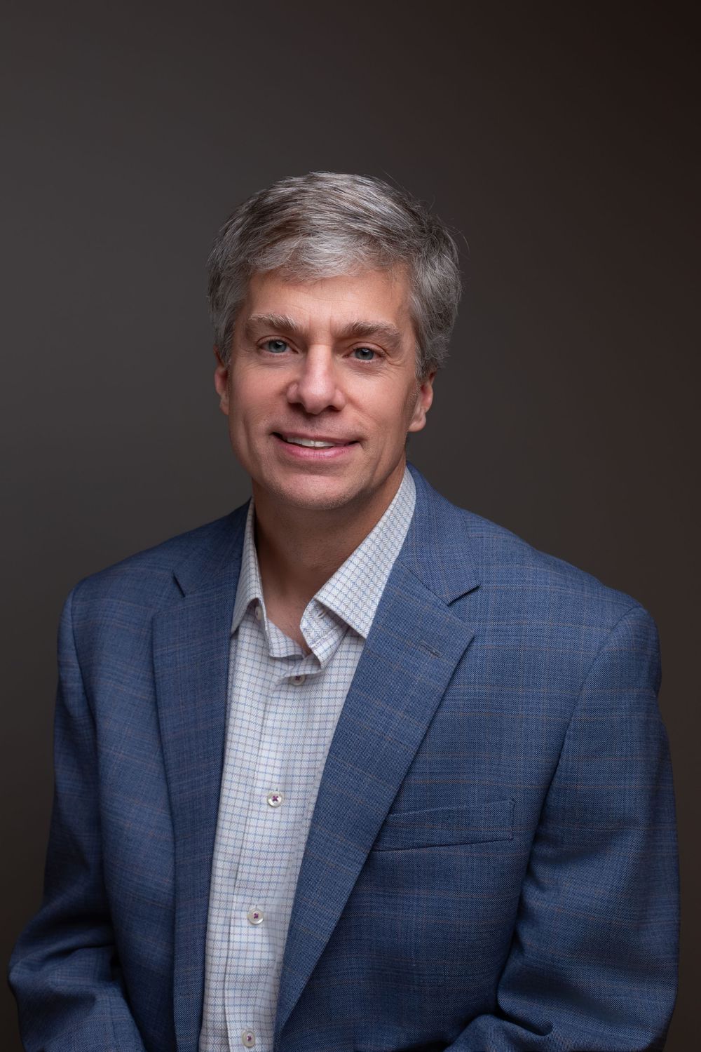 Professional headshot of a middle-aged man with gray hair wearing a blue blazer and checkered shirt, smiling confidently.