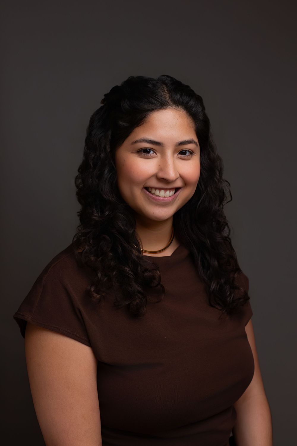 Smiling young woman with curly dark hair wearing a brown top, posing for a professional headshot against a gray background.