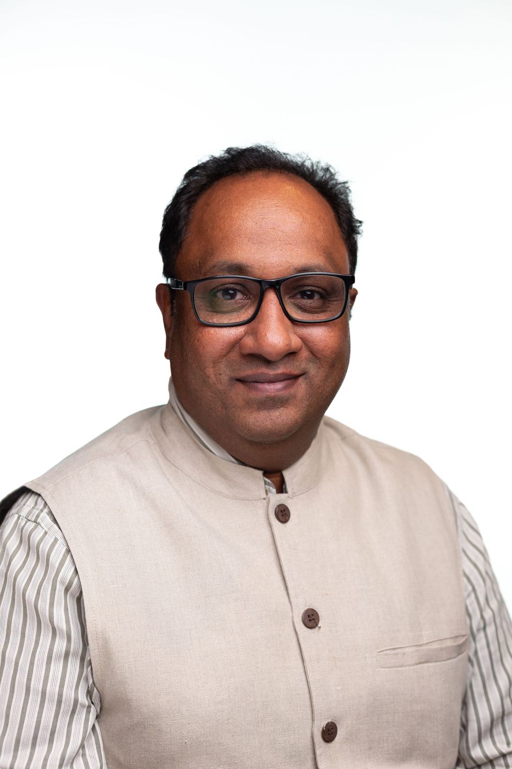 Professional portrait of person wearing glasses and a light-colored Nehru-style vest over striped shirt against white background.