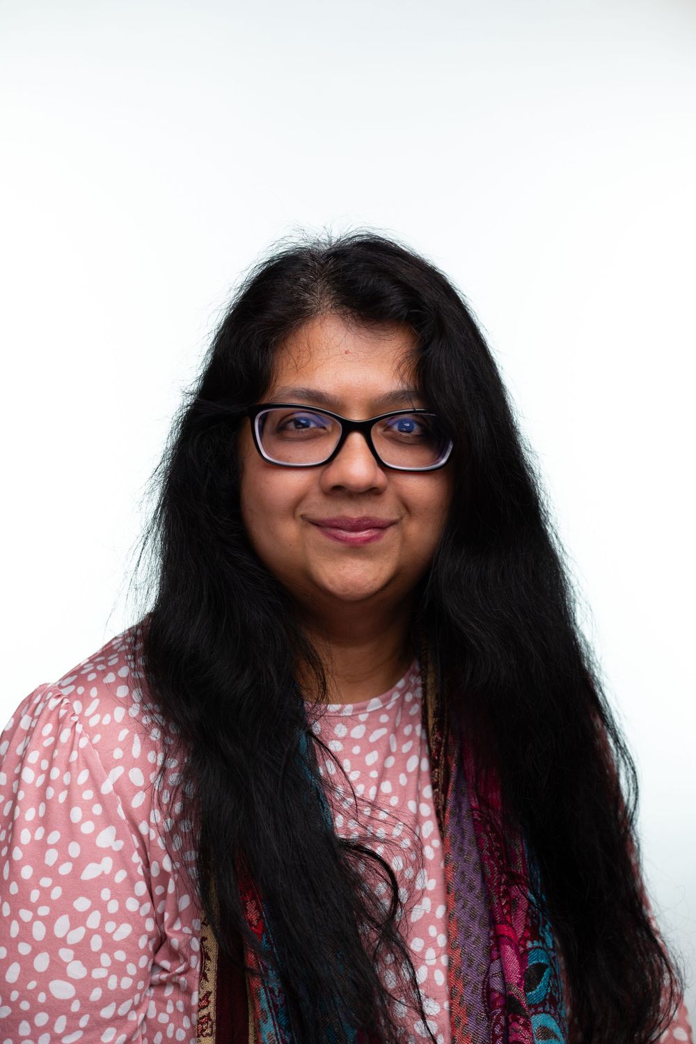 Portrait of person with long dark hair wearing glasses and pink patterned top against white background.