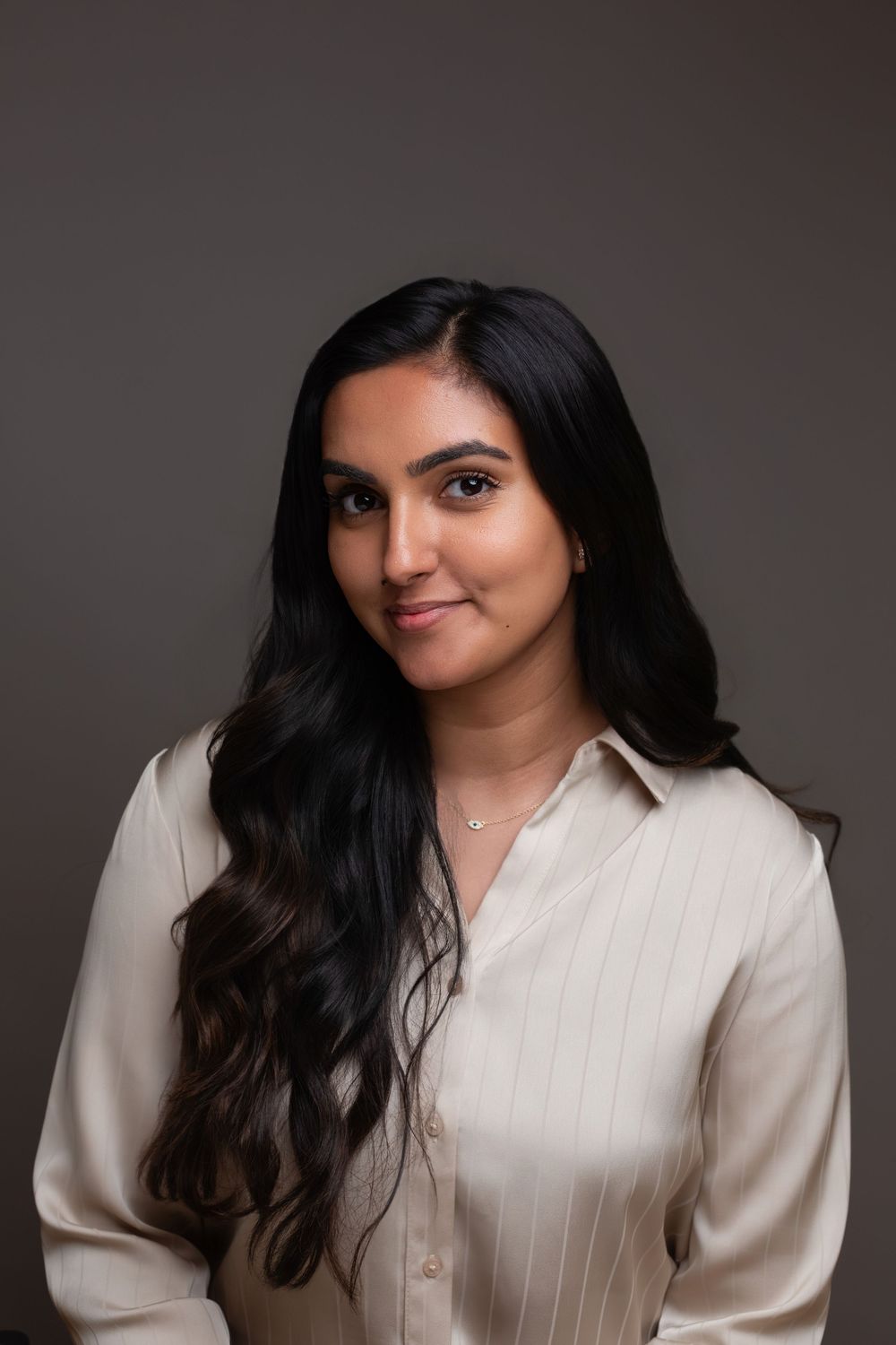 Young woman with long dark hair wearing a cream satin blouse and delicate necklace, posing against a gray background.