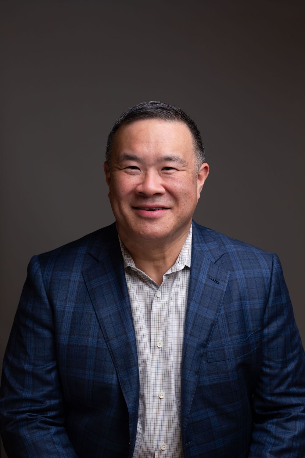 Professional headshot of a smiling middle-aged man in a blue plaid blazer against a gray background.