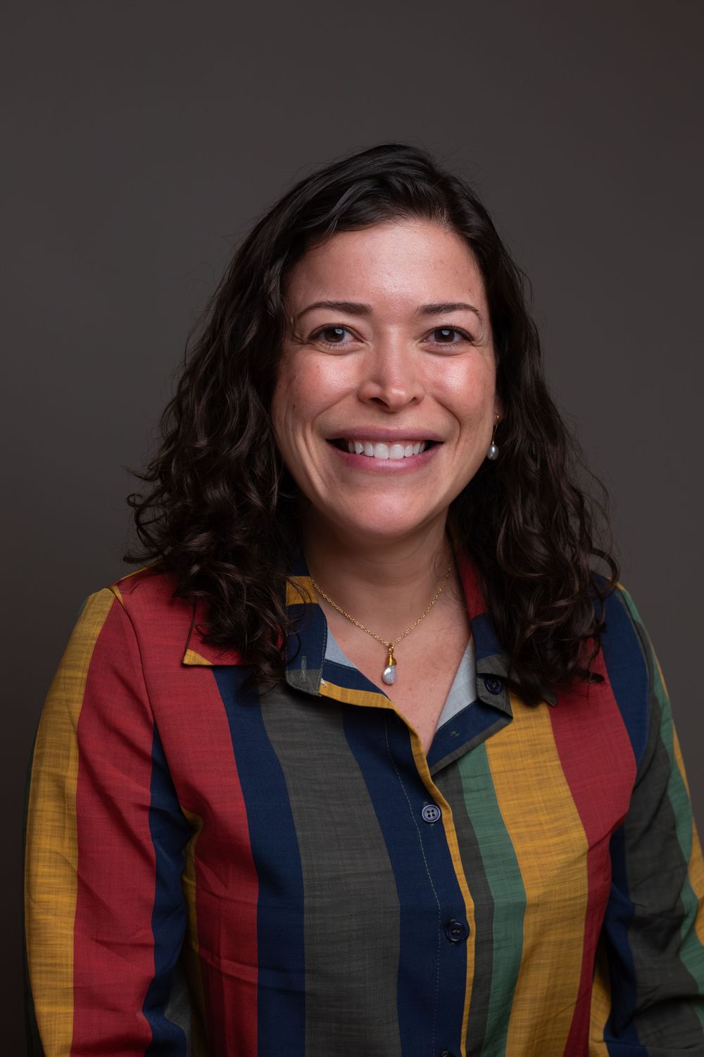 Smiling woman with curly dark hair wearing a colorful striped shirt, professional headshot on gray background.