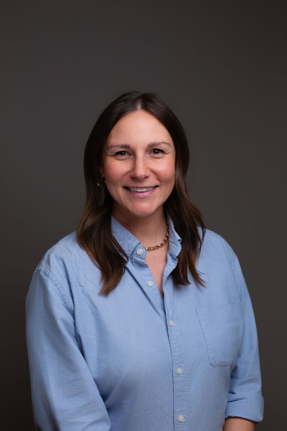 A smiling woman with brown hair wearing a light blue button-up shirt and gold necklace poses for a professional headshot.