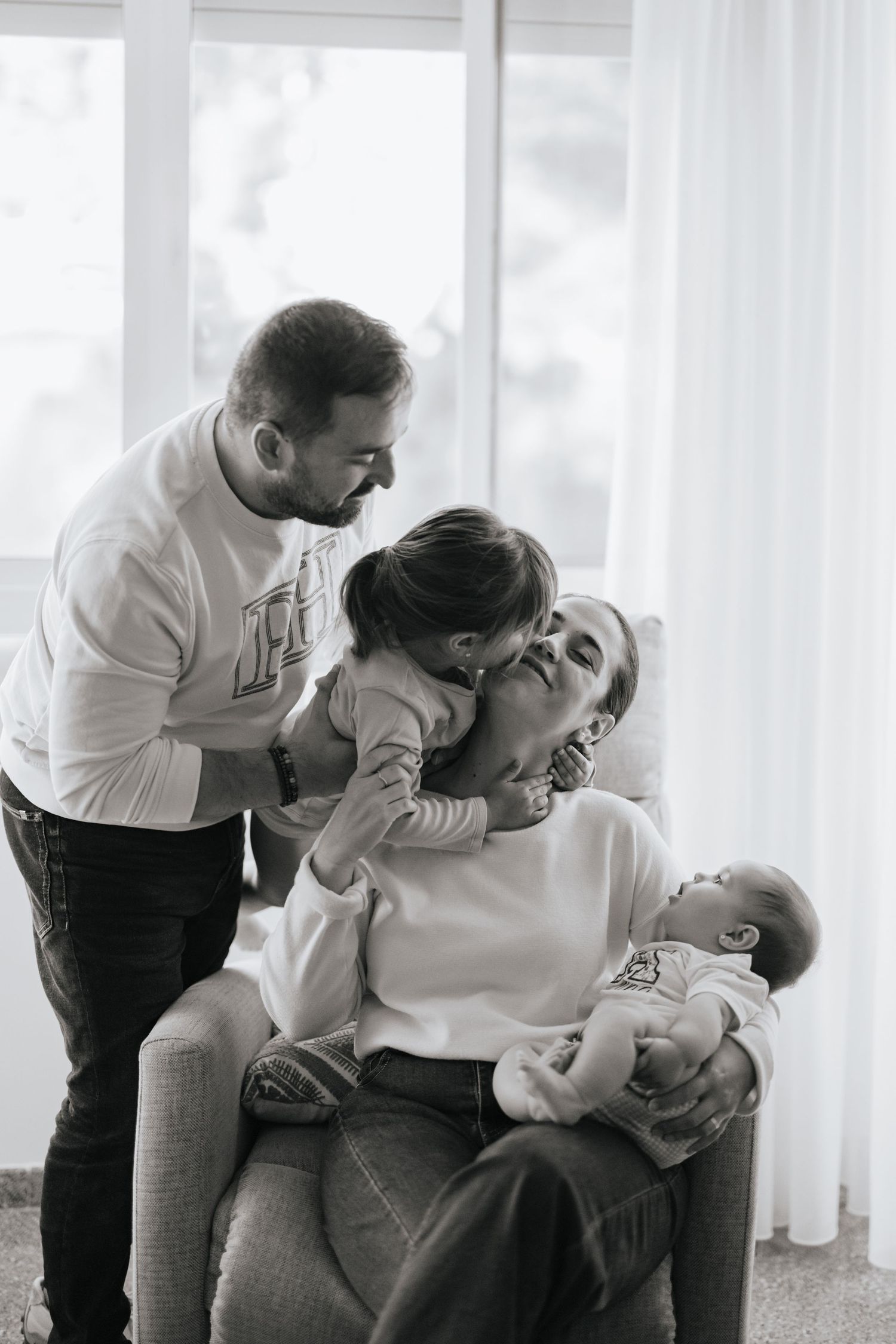 Black and white photo of a family sharing an intimate moment together by a window, wearing casual white tops and jeans.