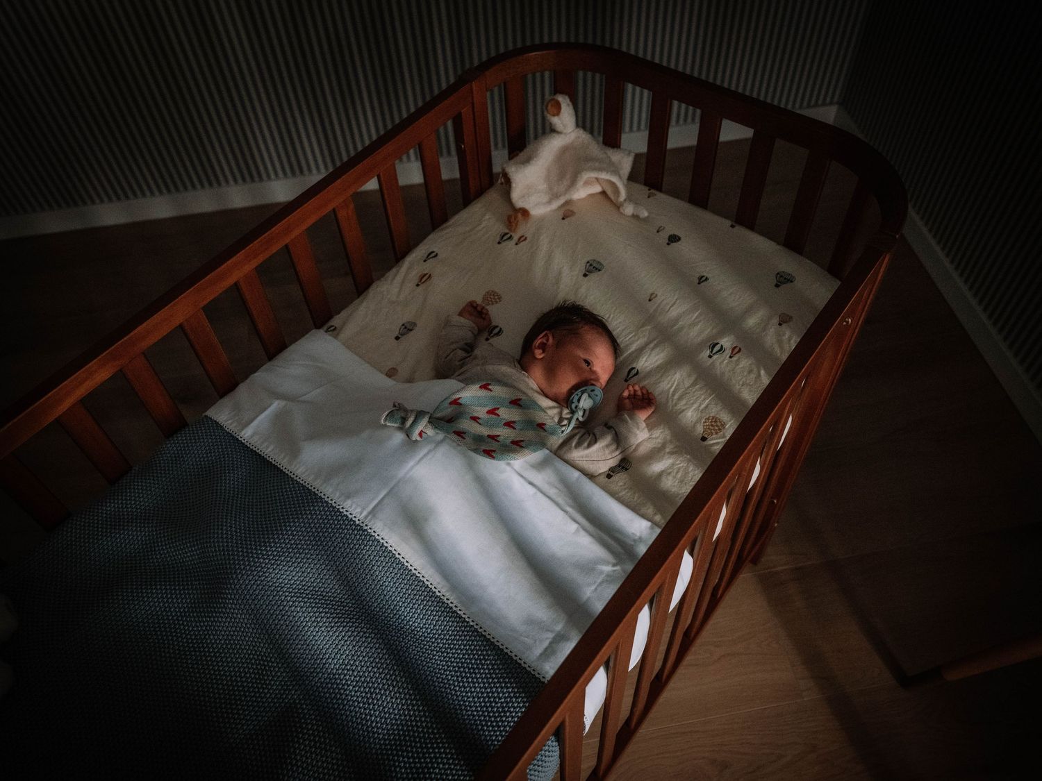Newborn baby sleeping peacefully in a wooden crib with white bedding in a dimly lit nursery room.