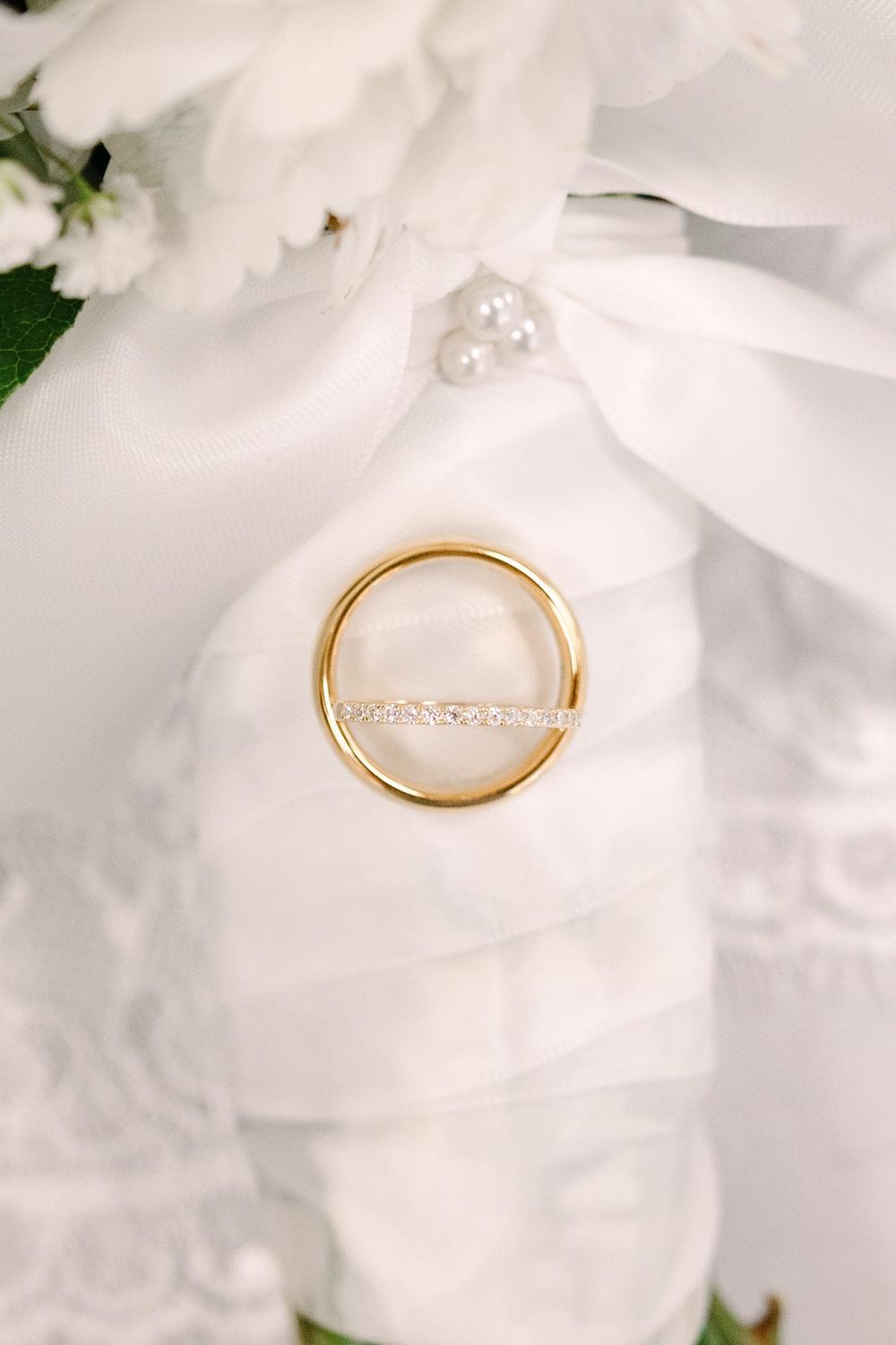A delicate gold ring with a diamond band rests on white flower petals in an elegant wedding photography composition.