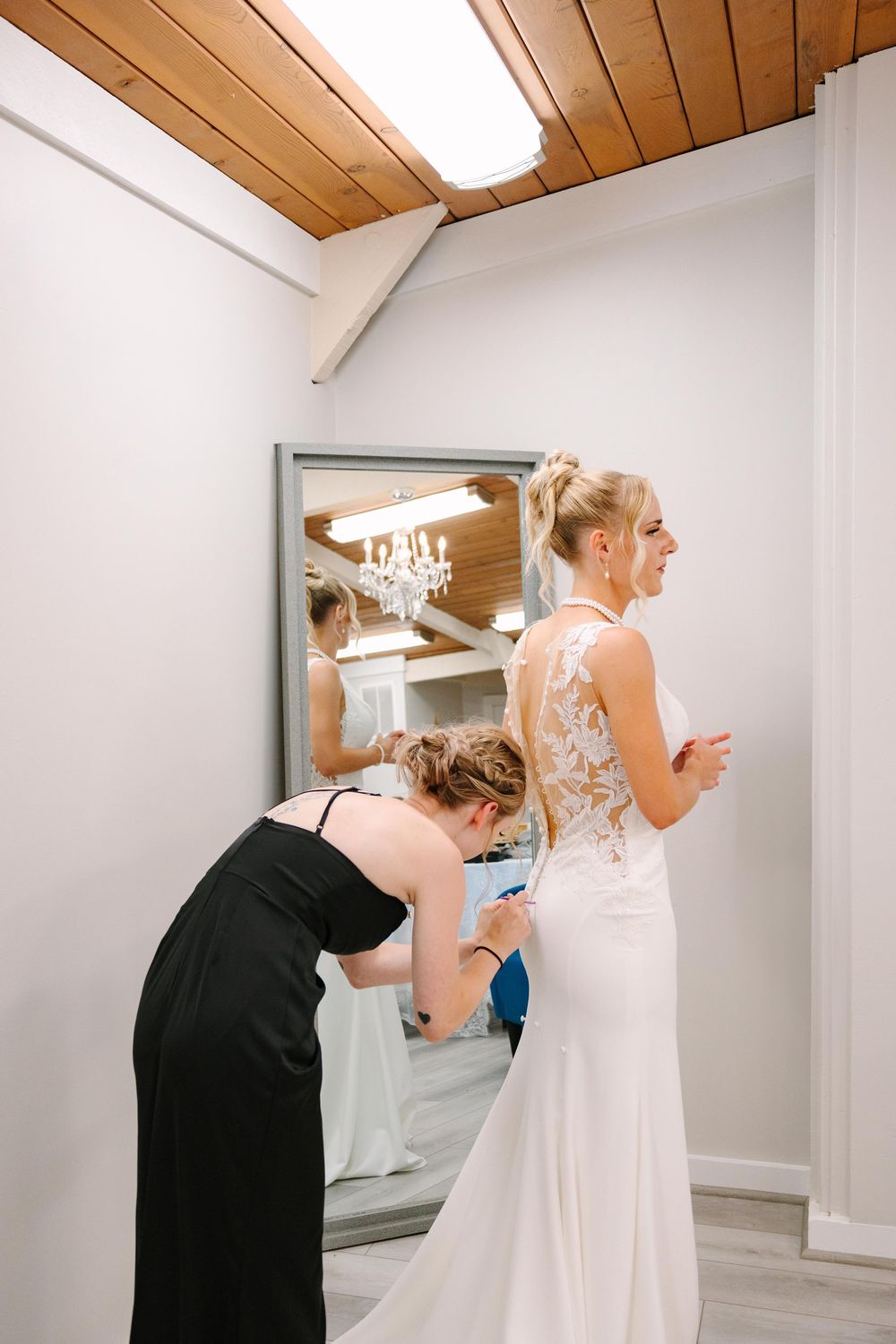 A woman in a white lace wedding dress being helped with final adjustments in front of a mirror.