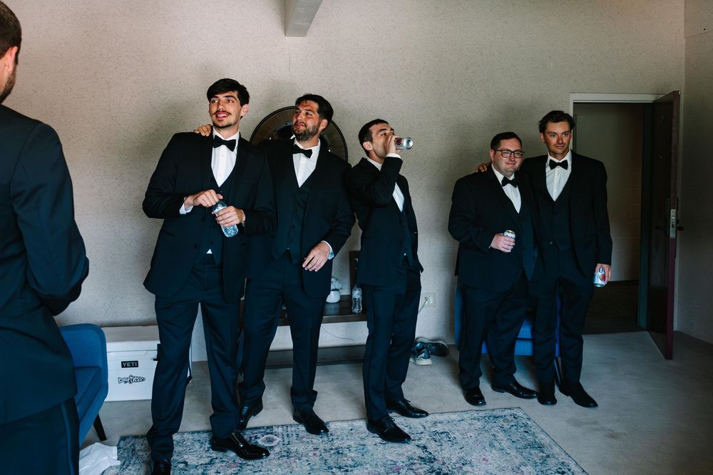 A group of groomsmen in black tuxedos and bow ties laugh together while standing in a hallway.